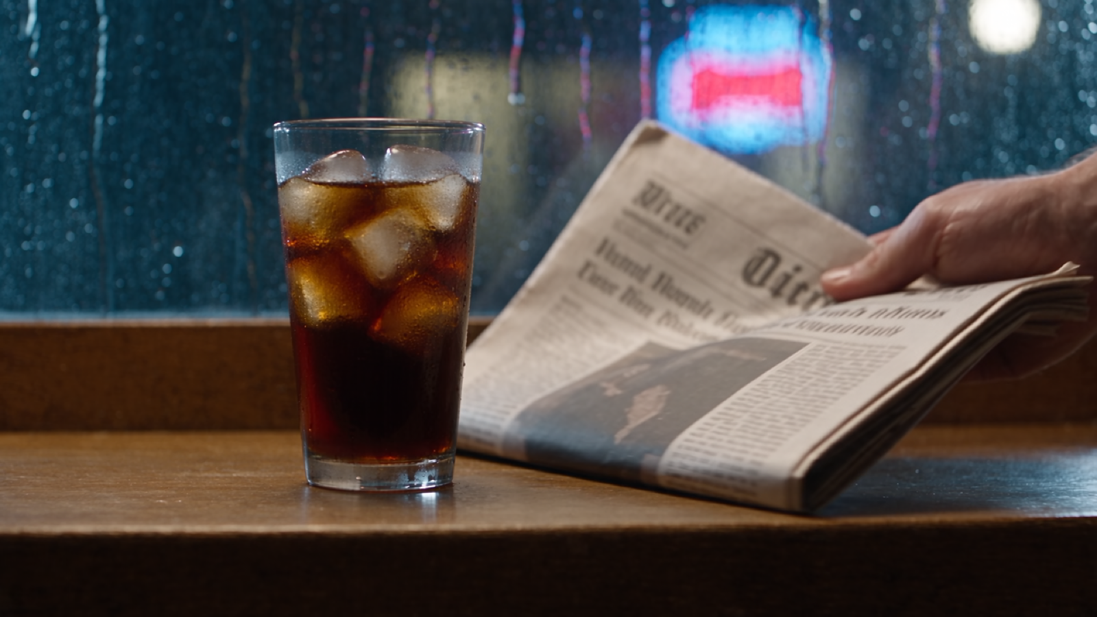 A frame from Seedance 2.5 at native 1080p: an iced coffee glass on a wooden cafe table, condensation beading on the sides, a hand resting on a folded newspaper, rain-streaked window and blurred neon behind